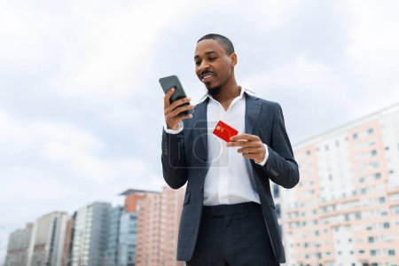 Téléchargez les photos : Online Banking. Black Businessman Using Smartphone And Credit Card Outdoors, Young African American Male Entrepreneur In Suit Enjoying E-Commerce, Standing Over Urban Buildings Background, Copy Space - en image libre de droit