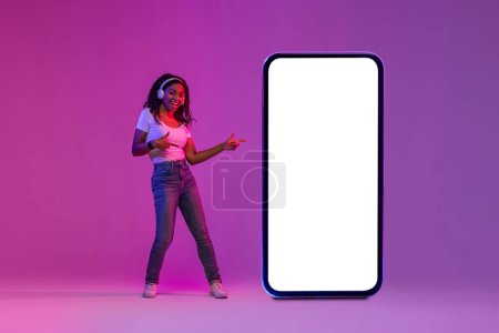 Téléchargez les photos : Check This. Cheerful Black Female Pointing At Big Blank Smartphone With White Screen While Standing In Neon Light Over Purple Gradient Background, Happy Lady Recommending New App, Collage, Mockup - en image libre de droit