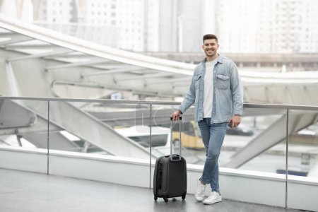 Photo for Cheerful Tourist Guy Standing With Travel Suitcase Smiling To Camera Posing At Airport Or Modern Train Station. Vacation And Tourism Concept. Full Length Shot, Copy Space - Royalty Free Image