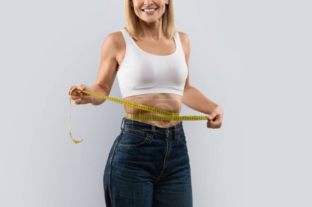Foto de Cropped of slim blonde woman in white top and dark jeans measuring her waist with measure tape, isolated on grey studio background, copy space. Weight loss, slimming concept - Imagen libre de derechos
