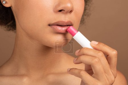 Téléchargez les photos : Serious young black curly lady with perfect skin applies lipstick on lips for moisturizing isolated on brown background, studio, cropped. Ad and offer of cosmetics treatment, beauty care, procedures - en image libre de droit