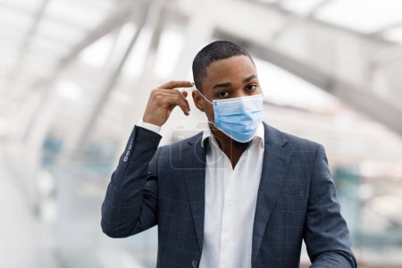 Téléchargez les photos : Young Black Businessman Taking On Medical Face Mask While Walking In Airport, African American Male Wearing Suit Following Protective Measures During Travel, Having Business Trip During Pandemic - en image libre de droit