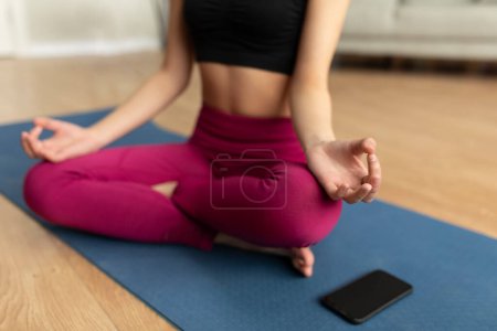 Foto de Sports, meditation, online training at home. Young lady practicing yoga in lotus position on mat with smartphone with blank screen, mockup - Imagen libre de derechos