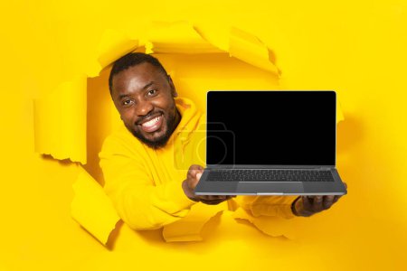 Foto de Hombre negro excitado sosteniendo el ordenador portátil con la pantalla vacía en blanco que muestra el dispositivo a través del agujero roto de papel amarillo, maqueta. Gadget con espacio de copia para plantilla de sitio web - Imagen libre de derechos