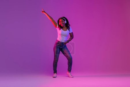 Téléchargez les photos : Joyful Black Woman Wearing Wireless Headphones Having Fun In Neon Light Over Gradient Purple Background, Happy African American Female Listening Favorite Music And Dancing, Full Length Shot - en image libre de droit