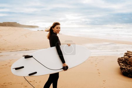 Deportes extremos. Mujer joven en traje de baño caminando en la playa con tabla de surf, mirando y sonriendo a la cámara, junto al mar en el fondo, espacio de copia gratis
