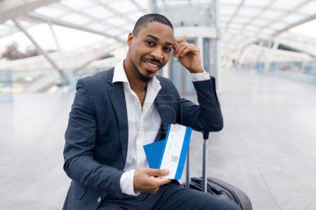 Téléchargez les photos : Portrait Of Handsome Young Black Businessman With Passport And Flight Tickets In Hand Waiting For Flight At Airport, Smiling African American Male Wearing Suit Sitting With Luggage At Terminal Hall - en image libre de droit
