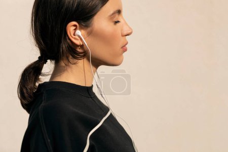 Foto de Calm caucasian millennial woman in sportswear and headphones with closed eyes enjoys fitness and jogging outdoors on white wall background. Sports and music, weight loss, active, body care and workout - Imagen libre de derechos