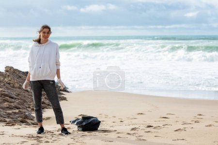 Foto de Young female activist standing with plastic bag on the beach by seaside, free copy space. Cleaning of the coastal zone and wild beaches. The concept of Earth Day - Imagen libre de derechos