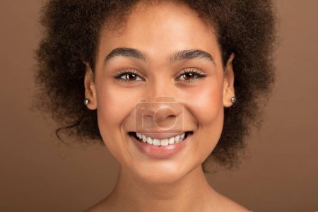 Téléchargez les photos : Portrait de jeune femme bouclée noire souriante avec une peau et des dents parfaites, maquillage nu regardant la caméra, isolé sur fond brun, studio, gros plan. Traitement de la peau, soins de beauté, procédures de spa - en image libre de droit