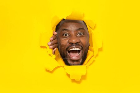 Photo for Wow. Emotional black man with open mouth breaking and looking through hole in slit yellow paper background. African american male feeling amazed at big sale or great news - Royalty Free Image