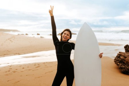 Photo for Young fit woman standing with surfboard on beach by seaside, smiling at camera and gesturing peace sign, enjoying active recreation, free space - Royalty Free Image