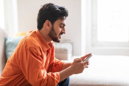 Téléchargez les photos : Heureux jeune arabe barbu attrayant en chemise orange refroidissant à la maison, assis sur le canapé dans le salon confortable, en utilisant un smartphone flambant neuf, bavarder avec les dames sur sortir ensemble app, espace de copie, vue latérale - en image libre de droit