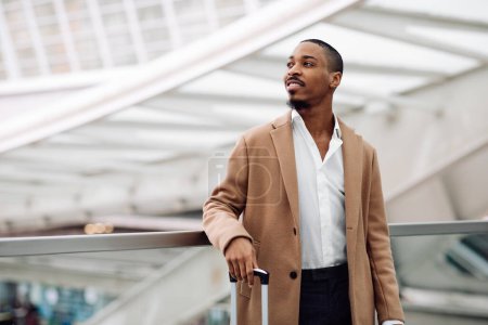 Photo for Handsome Black Man Wearing Stylish Coat Standing At Modern Airport, Portrait Of Young African American Male With Suitcase Waiting For His Flight At Terminal Hall, Ready For Air Travel, Copy Space - Royalty Free Image