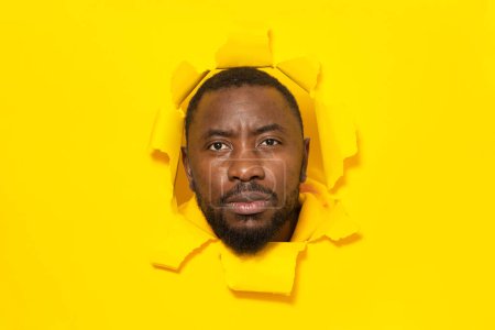 Téléchargez les photos : Sérieux homme afro-américain posant en papier jaune déchiré regardant la caméra à travers le trou, gros plan, espace libre - en image libre de droit