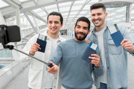 Foto de Travel Blog. Three Male Travelers Taking Photo On Phone Or Making Video Traveling Abroad In Modern Airport Indoors. Happy Friends Going On Vacation And Having Fun Waiting For Flight - Imagen libre de derechos