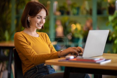 Mujer joven morena bonita feliz en traje informal freelancer sentado en la mesa delante de la computadora portátil, escribiendo en el teclado de la computadora, utilizando auriculares, trabajando desde la cafetería, vista lateral, espacio de copia