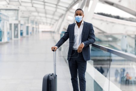 Photo for Young black businessman wearing suit and protective medical mask standing on walkway at airport terminal, african american male waiting for flight boarding, having business travel during pandemic - Royalty Free Image