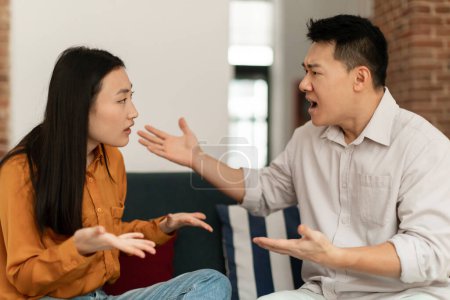 Photo for Angry asian spouses having quarrel experiencing crisis in relationship, sitting together on sofa at home. Middle aged man and yougn woman having conflict breaking up - Royalty Free Image