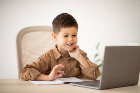Photo for Elementary education remotely. Smiling caucasian little child sit at table, make notes, watch lesson on computer in room interior. Study at school, kindergarten and home, childhood and video call - Royalty Free Image