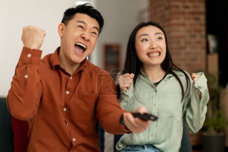 Foto de Emotional japanese spouses shaking fists and shouting while watching sport channel on TV, sitting on sofa at home. Sport and television programming, weekend leisure - Imagen libre de derechos