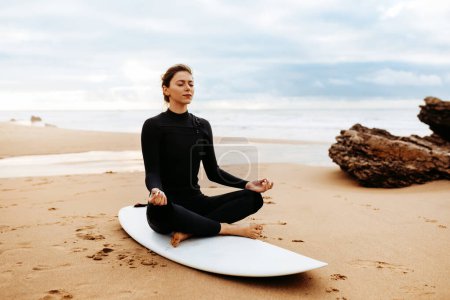 Téléchargez les photos : Planche de yoga. Fit femme européenne en maillot de bain méditant sur la planche de surf sur la plage au bord de la mer, assis les yeux fermés dans la pose de lotus, espace libre. Relaxation et cal - en image libre de droit