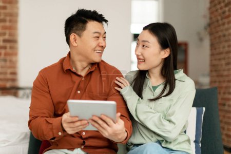 Foto de Happy asian lovers middle aged man and young woman sitting on sofa and using digital tablet, looking at each other and smiling, enjoying newest mobile application, home interior, copy space - Imagen libre de derechos