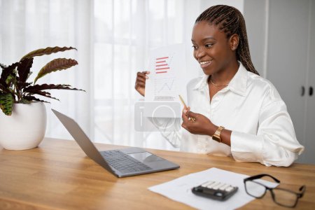 Cheerful attractive young black woman in smart casual sitting at workdesk at office, using laptop, showing papers at computer camera, have online business meeting with business partners, copy space