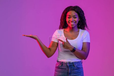 Téléchargez les photos : Check This. Smiling Young Black Woman Pointing At Her Open Empty Palm, Cheerful African American Female Recommending Invisible Object While Standing In Neon Light Over Purple Background, Copy Space - en image libre de droit