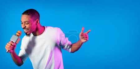 Photo for Cheerful Black Millennial Guy Holding Microphone And Singing, Young Joyful African American Hipster Man Enjoying Karaoke, Having Fun While Standing On Blue Studio Background In Neon Light, Copy Space - Royalty Free Image