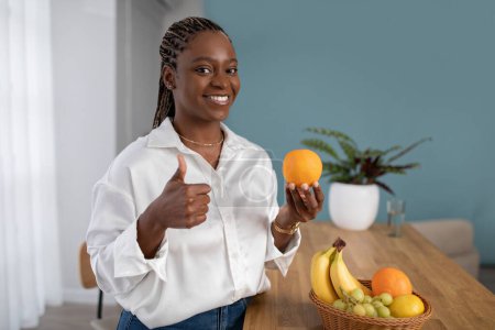 Photo for Happy attractive young black woman in smart casual outfit nutritionist showing fresh juicy orange and thumb up, smiling at camera, recommending food full of vitamins and minerals, copy space - Royalty Free Image