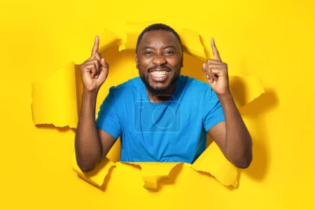 Téléchargez les photos : Portrait d'un homme afro-américain excité pointant les doigts vers le haut et souriant, debout dans un trou de papier déchiré, posant à travers une percée de fond jaune - en image libre de droit