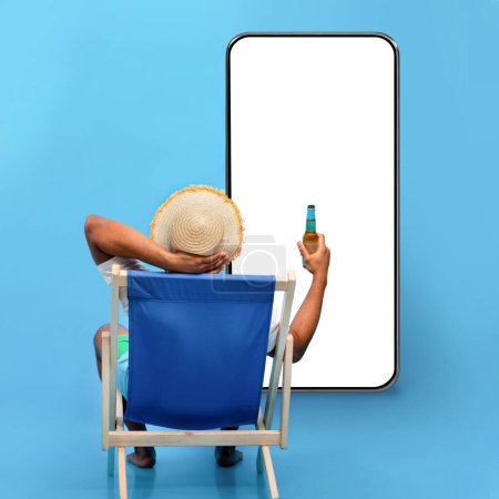 Téléchargez les photos : Back view of young black guy sitting in lounge chair with bottle of beer, sipping alcoholic beverage, looking at phone with blank screen on blue studio background, mockup for travelling offer, collage - en image libre de droit
