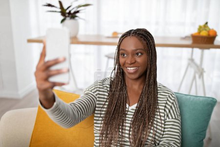 Téléchargez les photos : Positif souriant jolie jeune femme noire avec de longues tresses assises sur le canapé à la maison, prenant selfie sur téléphone portable, se refroidissant dans le salon élégant confortable, blogueuse publiant du contenu sur les médias sociaux - en image libre de droit
