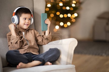 Téléchargez les photos : Glad european small kid in wireless headphones calls by phone, sits in armchair in living room interior. App for meeting remotely, entertainment, fun and communication, education with device at home - en image libre de droit