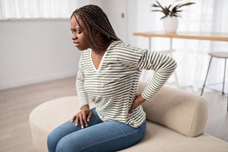 Photo for Upset young black long-haired woman in pain sitting on couch, touching her lower back, african american lady suffering from backache, muscle strain, period cramps, copy space - Royalty Free Image
