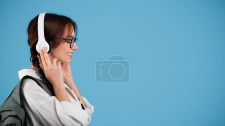 Téléchargez les photos : Smiling caucasian teen girl with pigtails in glasses with headphones, student with backpack listens to music, enjoy relax, isolated on blue background, studio. Audio app for learn, study, education - en image libre de droit