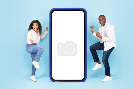 Téléchargez les photos : Overjoyed black spouses shaking clenched fists near big smartphone with empty white screen, celebrating win over blue studio background, mockup, full body length - en image libre de droit