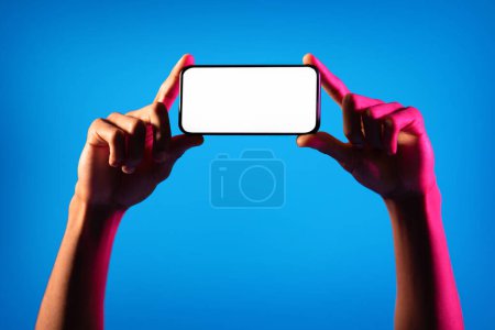 Photo for Online offer, mobile app. Cropped of african american man hands hand holding horizontal cell phone with white blank screen on blue studio background in neon light, mockup for advertisement, copy space - Royalty Free Image