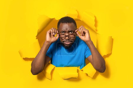 Foto de Angry african american man looking at camera through eyeglasses, noticing something, breaking through torn hole in yellow paper background, free space - Imagen libre de derechos