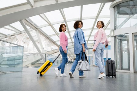 Foto de Tres hermosas mujeres caminando con equipaje en el aeropuerto y girando en la cámara, grupo de amigas jóvenes felices que viajan juntas al extranjero, señoras alegres posando en la terminal, espacio de copia - Imagen libre de derechos