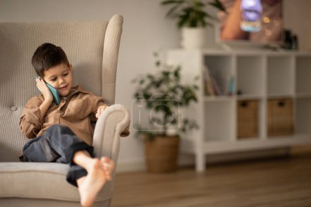 Foto de Meeting remotely, childhood. Glad european small kid calling on phone, sitting in armchair in living room interior, empty space. Fun, entertainment and communication, conversation with device at home - Imagen libre de derechos
