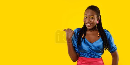 Téléchargez les photos : Cheerful African American Lady Pointing Thumb Finger Aside At Free Space Advertising Your Text Posing Over Yellow Background In Studio, Smiling To Camera. Check This Offer. Panorama - en image libre de droit