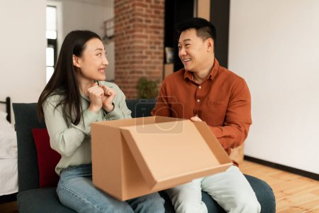 Téléchargez les photos : Bon concept de service de livraison. Étonnés conjoints asiatiques ouvrant la boîte de livraison à la maison, se regardant et souriant, montrant étonnement, intérieur de la maison, espace libre - en image libre de droit