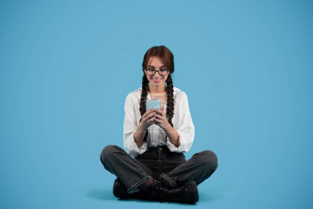 Téléchargez les photos : Smiling caucasian teen girl with pigtails student sit on floor, chatting on phone, isolated on blue background, studio. Communication, learn, knowledge and study with gadget, education ad and offer - en image libre de droit
