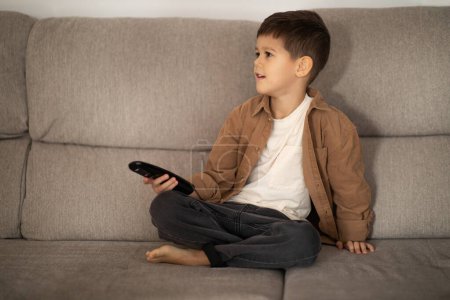 Foto de Serious cute european small kid uses remote control, switches channel on TV set, enjoy spare time alone, sits on sofa in living room, empty space. Education with device at home, cartoons and video - Imagen libre de derechos