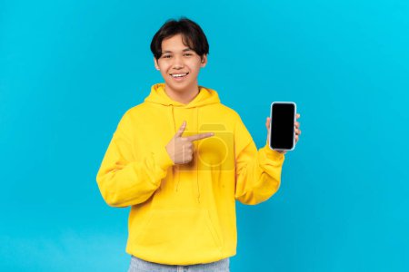 Téléchargez les photos : Cheerful Korean Teen Guy Holding Phone With Empty Screen And Pointing Finger Recommending Mobile Offer Or New Application Posing On Blue Studio Background. Technology And Gadgets. Mockup - en image libre de droit