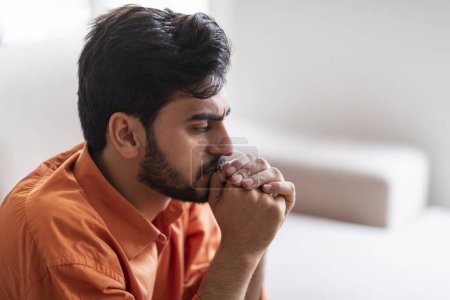 Photo for Mental health, depression concept. Frustrated thoughtful young bearded arab guy sitting on sofa alone at home, touching his mouth, looking down, have hard thoughts, copy space, side view, closeup - Royalty Free Image