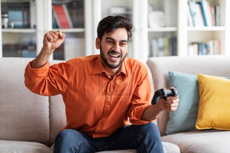 Photo for Cheerful happy young attractive arab guy in smart casual enjoying his new video game console at home, emotional man sitting on couch alone, holding joystick, gesturing and exclaiming, copy space - Royalty Free Image