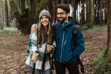 Téléchargez les photos : Happy young european male and woman tourists in jackets with backpack and trekking sticks walk in autumn forest, enjoy cold season, outdoor. Lifestyle, travel, adventure, nature and relationships - en image libre de droit